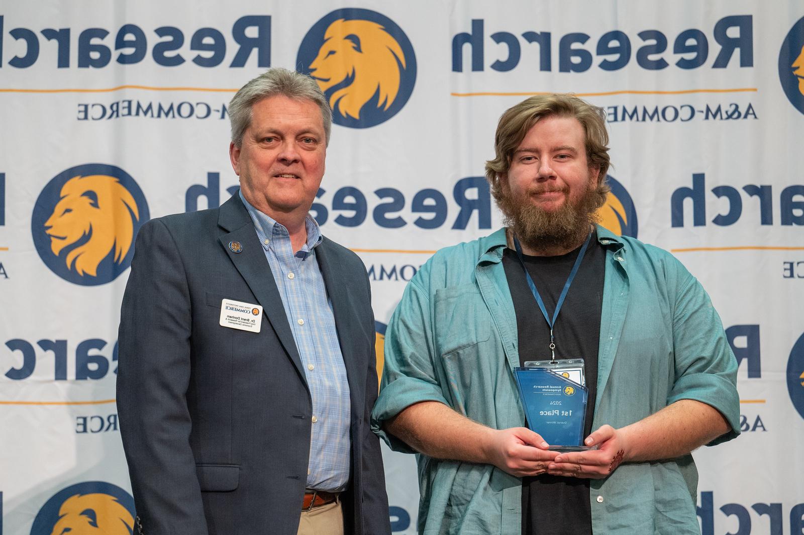 Joshua Belieu and Dr. Brent Donham stand in front of a white backdrop with A&M-Commerce Research logos, which include the university's lion head.